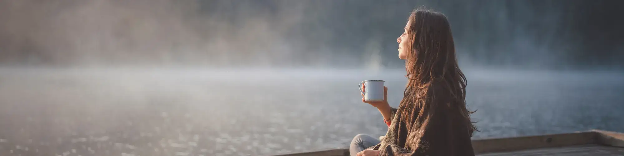 Frau sitzt entspannt am See mit einer Tasse in der Hand und atmet ruhig – Symbol für Achtsamkeit und innere Ruhe.