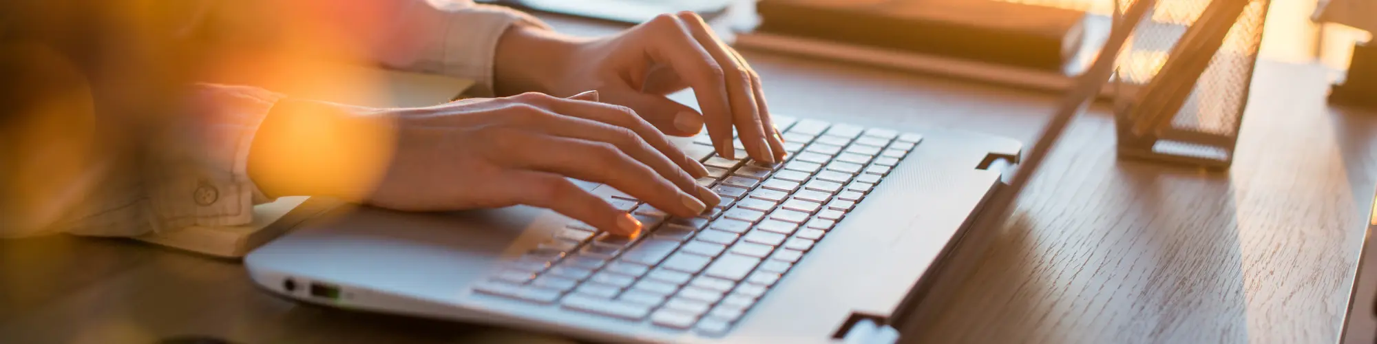 Hände tippen im warmen Sonnenlicht auf einer Laptop-Tastatur – ruhige Schreibszene.